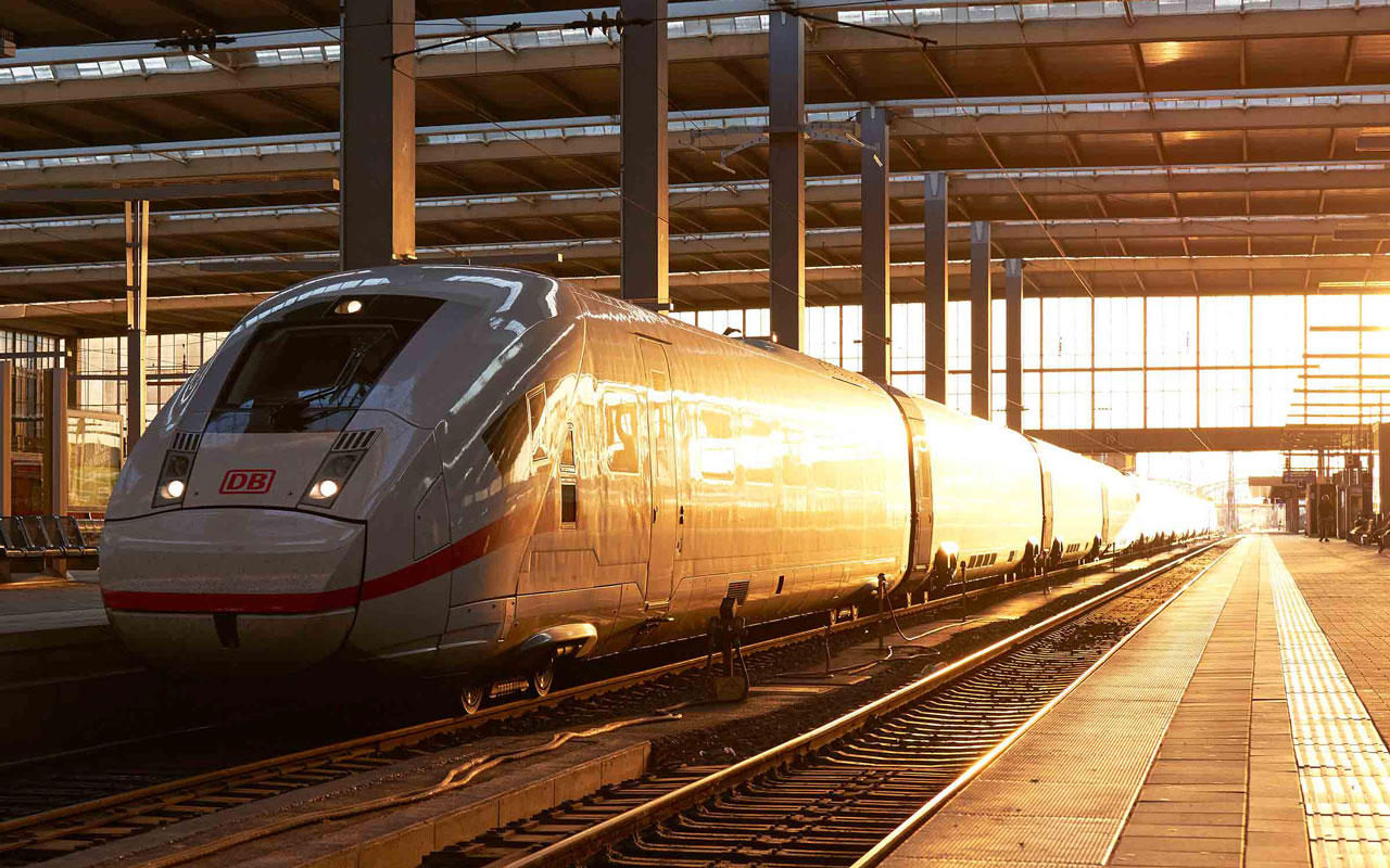 Schnellzug mit grauer und silberner Front und roten Linien in einer beleuchteten Bahnhalle, am Bahnsteig auf Gleisen stehend.