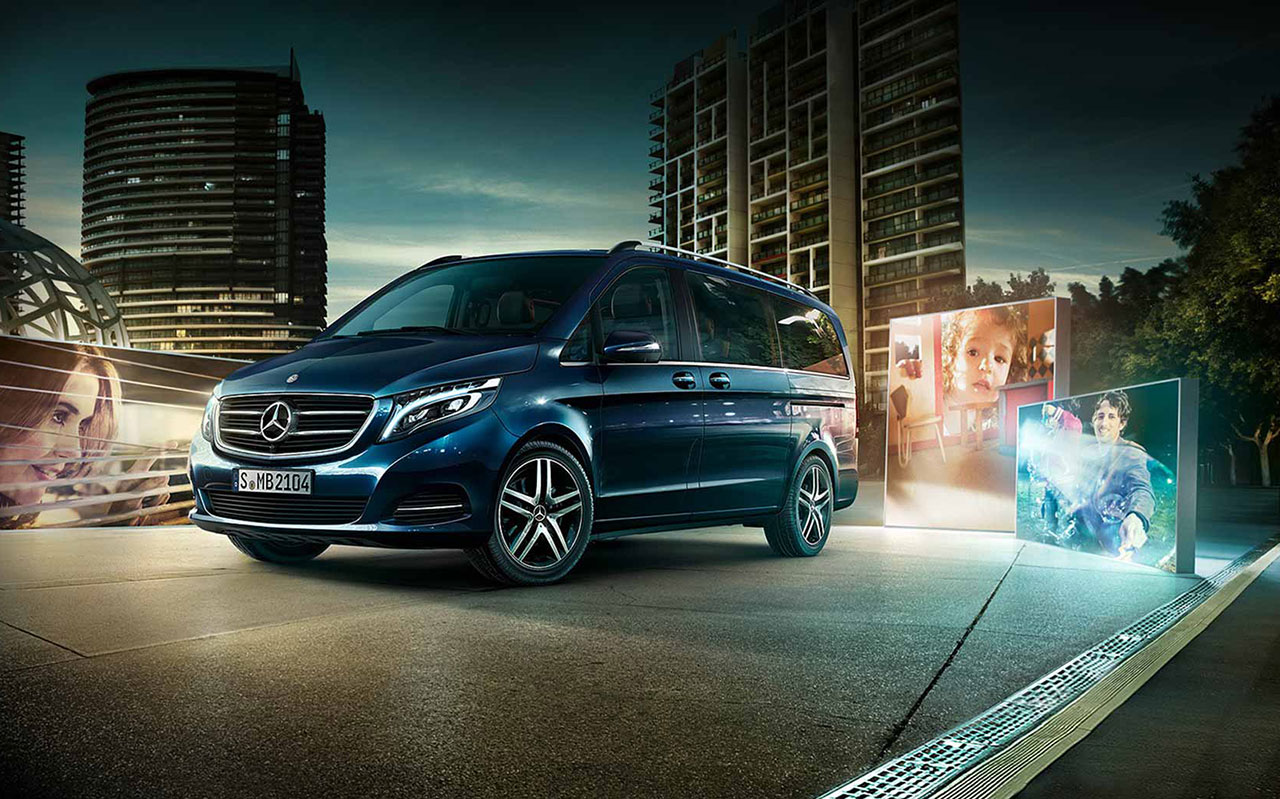 Dunkelblauer Mercedes-Minivan mit LED-Front und großen Felgen auf einer Stadtstraße vor Hochhäusern, mit rechten Werbedisplays und Bildmotiven im Hintergrund.