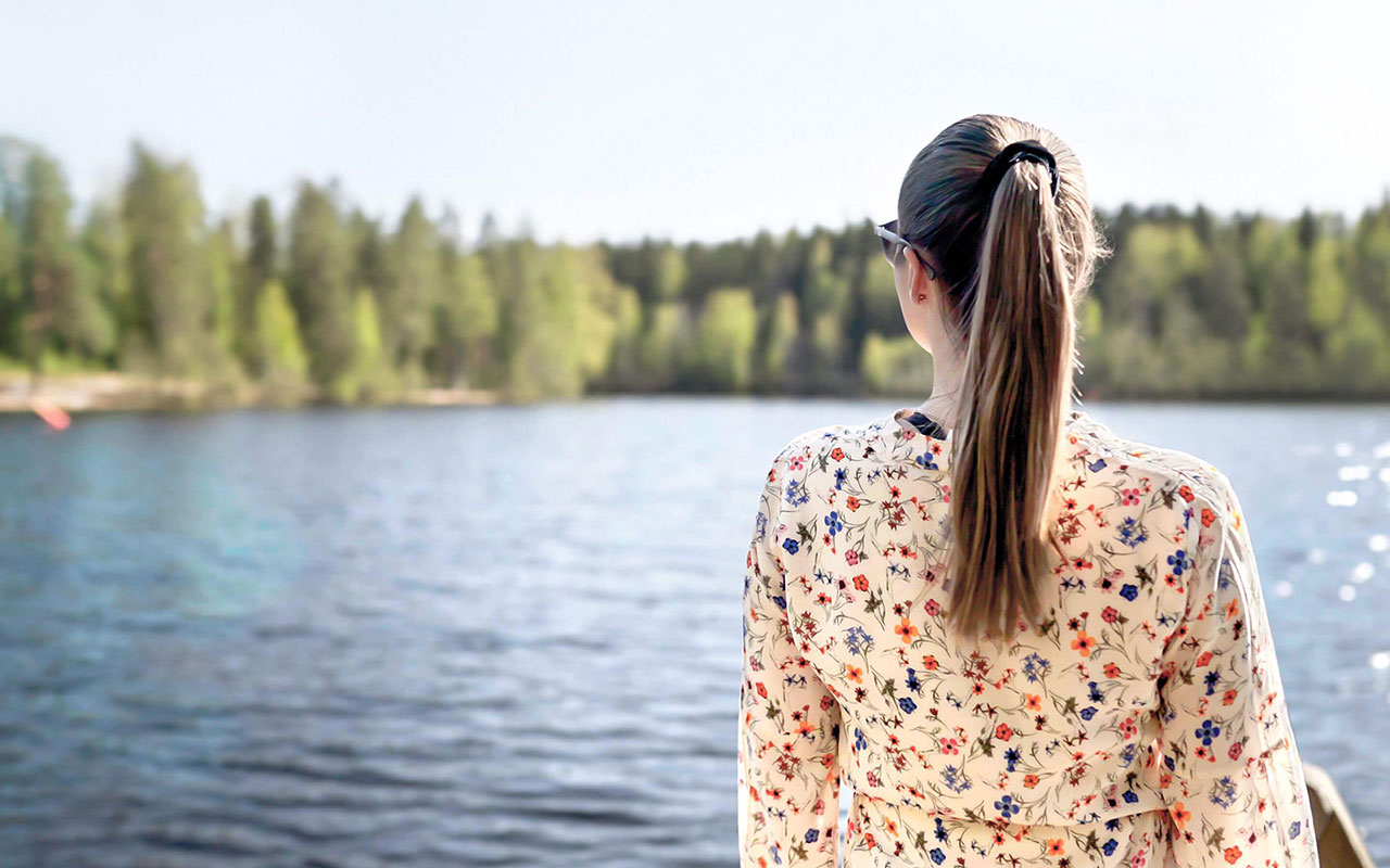 Person mit langem braunem Haar und Blumenbluse schaut an einem See nach links, im Hintergrund Wald und helles Ufer.