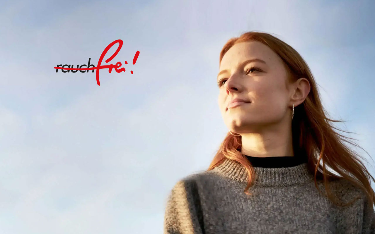 Frau im Halbporträt vor hellem Himmel, dezentes Lächeln, rotes-braunes Haar, grauer Strickpullover; links Textgrafik „rauchfrei“ und „Pf!“
