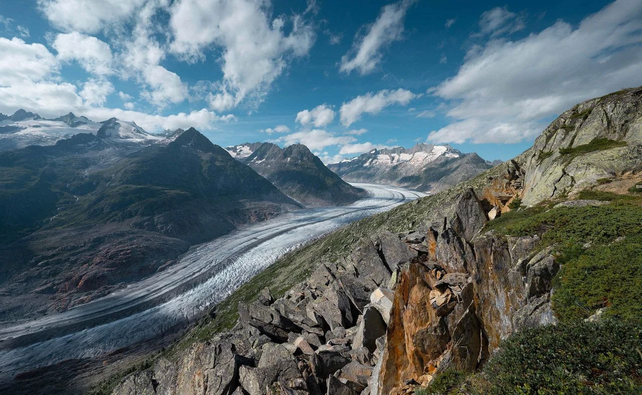 Panoramablick auf einen Gletscher im Tal mit steilen Felsbergen und blauem Himmel mit Wolken.